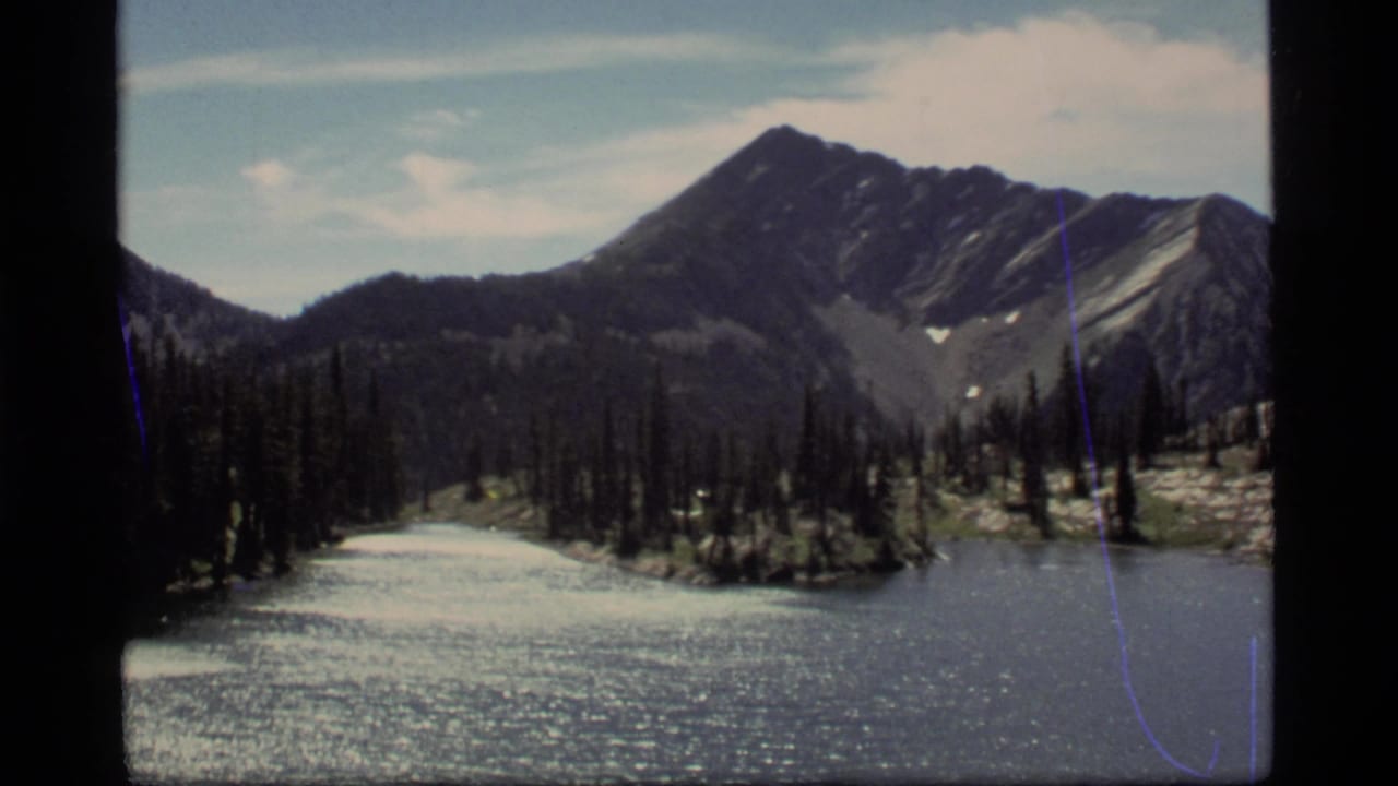 1977 8mm view of Sapphire Lake in Montana with dark conifers, bright water, and a rocky mountain ridge beyond.