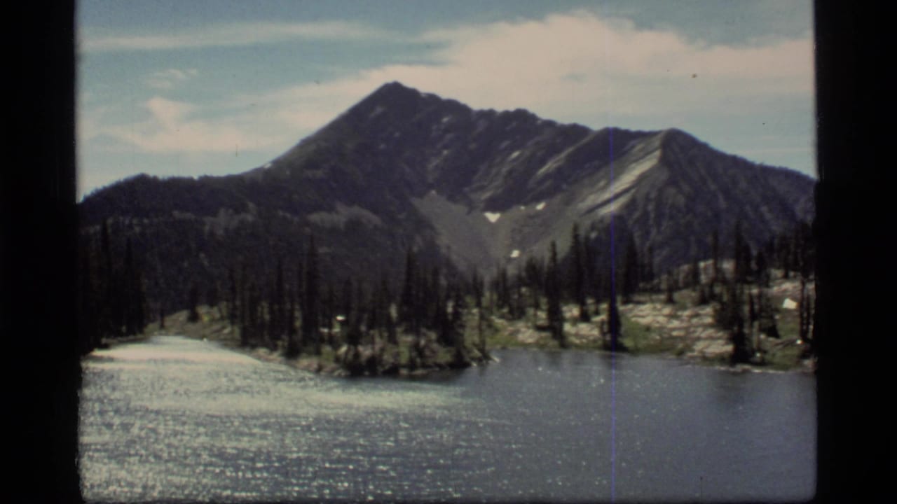 Mountain lake and forested shoreline below a dark ridgeline and pale clouds.