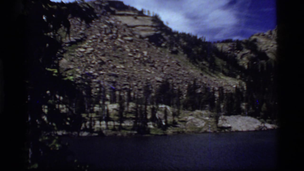 1977 8mm home-movie frame of Sapphire Lake, Montana, showing dark water, rocky slopes, evergreens, and blue sky.
