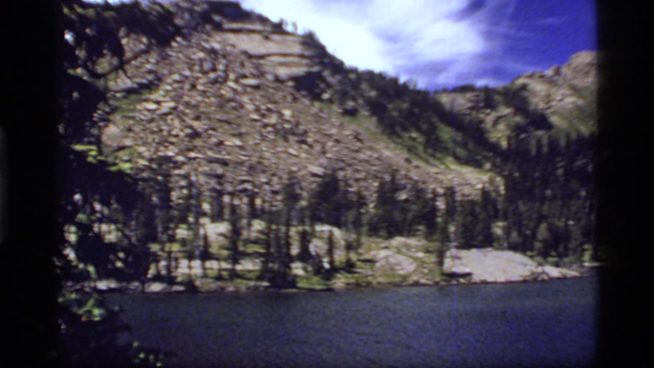 Rocky mountain slope and evergreen forest rising behind a dark lake under a blue sky.