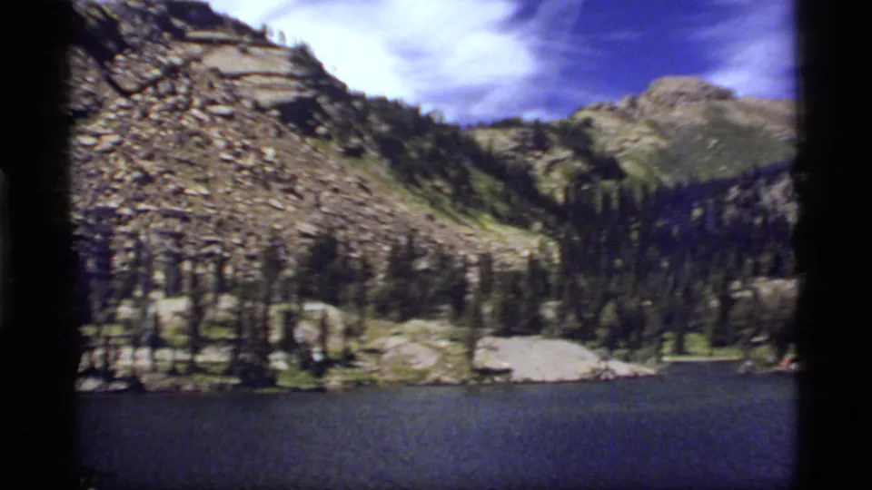 1977 8mm frame from Sapphire Lake, Montana, with water in the foreground and forested mountains under clouded blue sky.