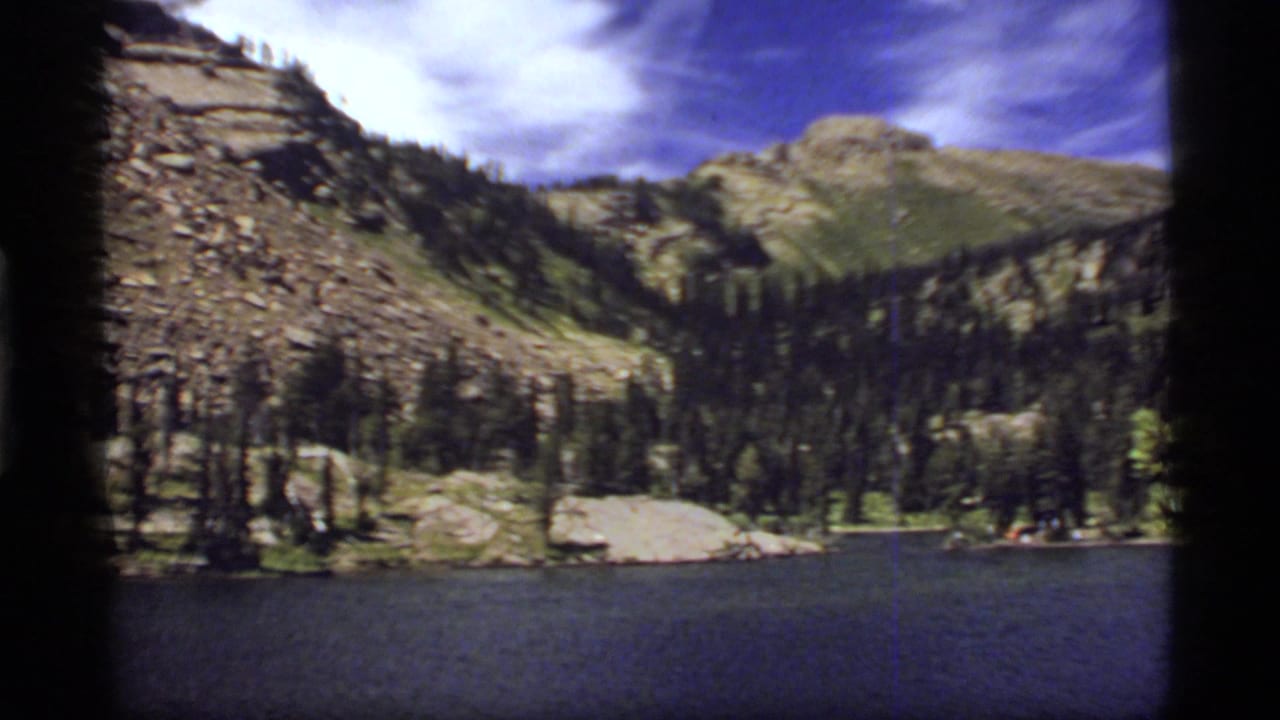 Mountain lake below rocky green slopes and pine forest under a blue sky.
