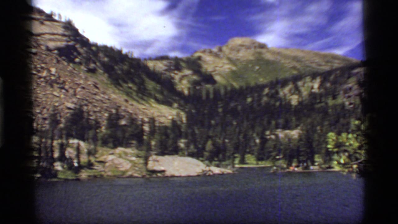 1977 8mm view across Sapphire Lake toward pine-covered shoreline, rocky mountains, and a partly cloudy blue sky.