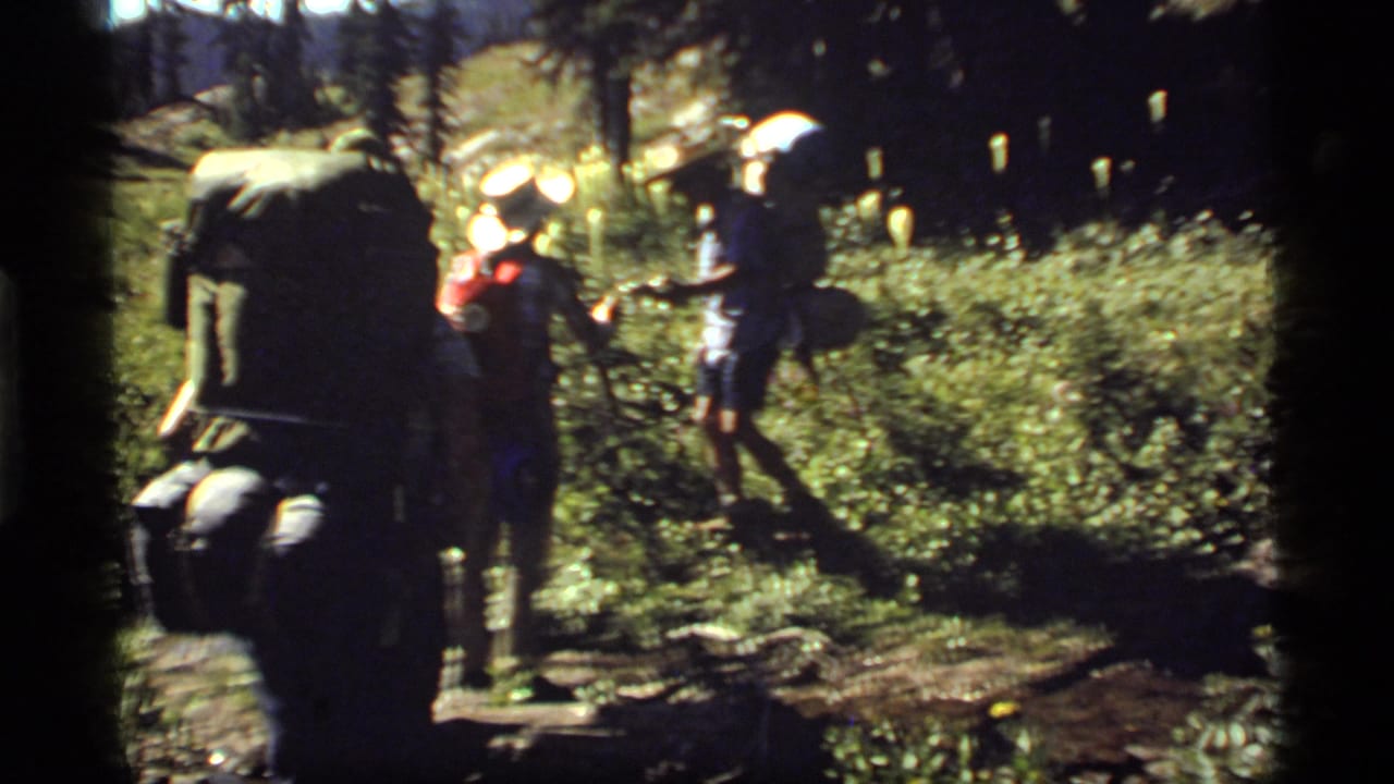 1977 Montana 8mm frame showing hikers with large packs moving through a shaded, rocky forest trail near Sapphire Lake.