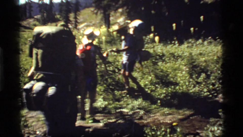 1977 Montana 8mm frame showing hikers with large packs moving through a shaded, rocky forest trail near Sapphire Lake.