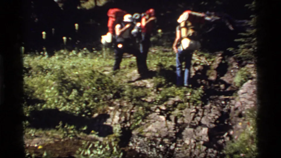 Two hikers with large packs stand near rocky ground and low vegetation in 1977 Sapphire Lake, Montana home-movie footage.