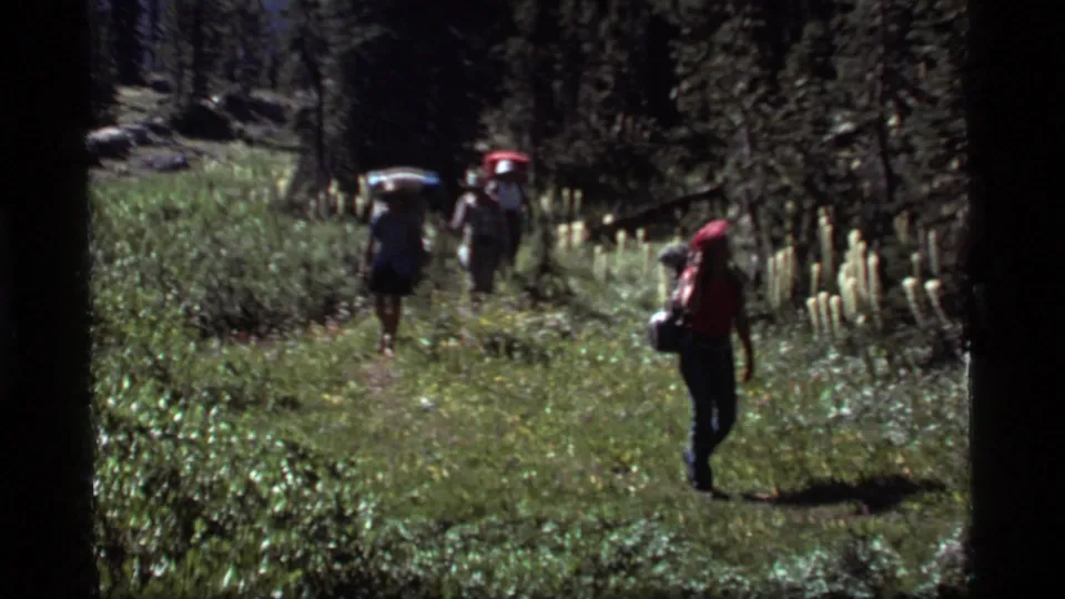 The 1977 Montana clip shows the group moving through a forest trail, with a red-shirted hiker closer to camera and backpackers ahead.
