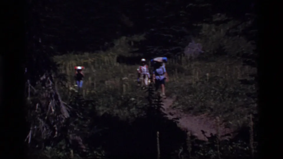 1977 8mm frame from Sapphire Lake, Montana, showing backpackers moving along a narrow sunlit forest trail.