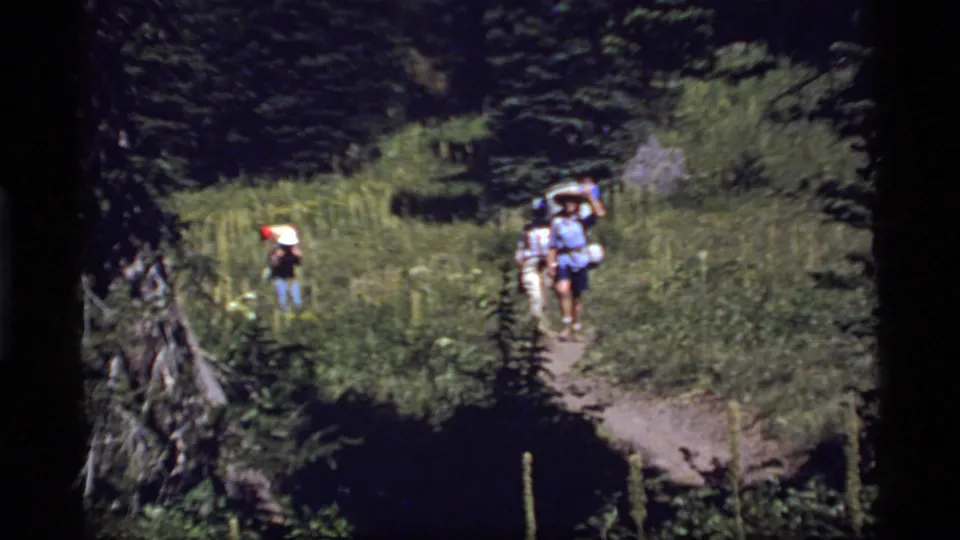 1977 Sapphire Lake, Montana home-movie frame of hikers with packs crossing a grassy forest trail opening.