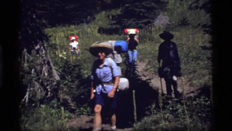 A hiker with a wide-brimmed hat and pack passes close to the camera on a 1977 Sapphire Lake, Montana forest trail.