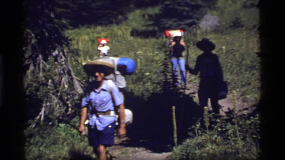 1977 Montana home-movie frame showing hikers with large packs and hats walking on a narrow forest trail.