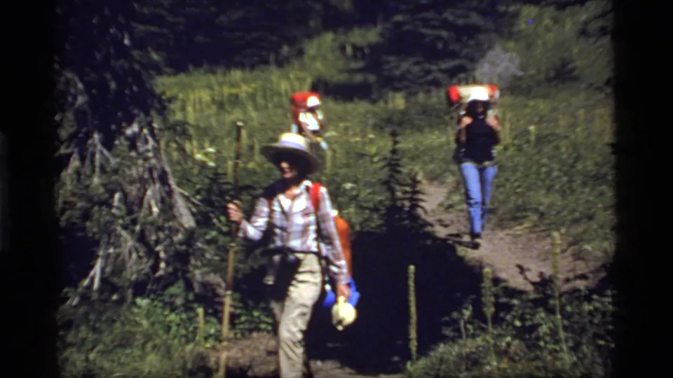 1977 Montana hiking footage shows an adult hiker with a pack and hat leading others along a wooded trail.