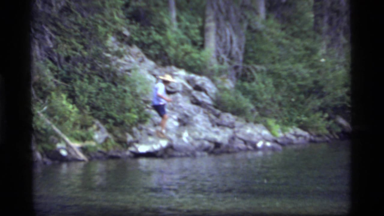 At Sapphire Lake, Montana in 1977, a man in shorts and a straw hat stands carefully on a rocky shoreline near dark water and trees.
