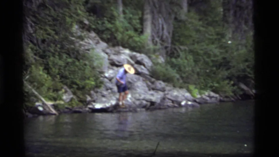 The 1977 Montana lake footage shows the man paused on the shoreline rocks, framed by shrubs, trees, and dark water.