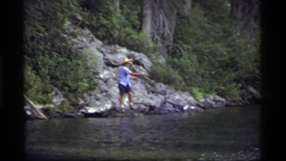 A man in shorts and a straw hat reaches toward the rocky bank at Sapphire Lake, with evergreen forest and water filling the frame.