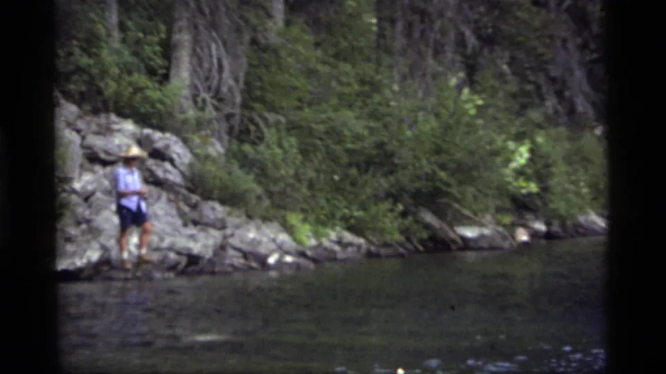 The fisher remains near the rocky bank, framed by dark water and dense forest in the 8mm Montana scene.