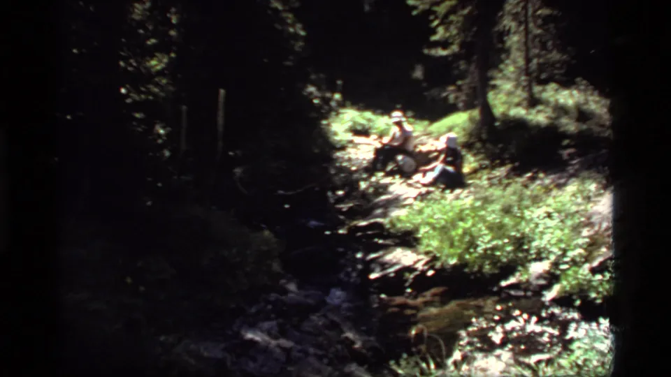 Two people appear seated beside a bright forest path or stream, continuing the 1977 Montana outdoor home-movie setting.