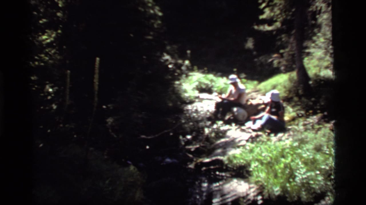 Two hikers rest beside a small stream in a wooded area near Sapphire Lake, Montana, in 1977 home-movie footage.