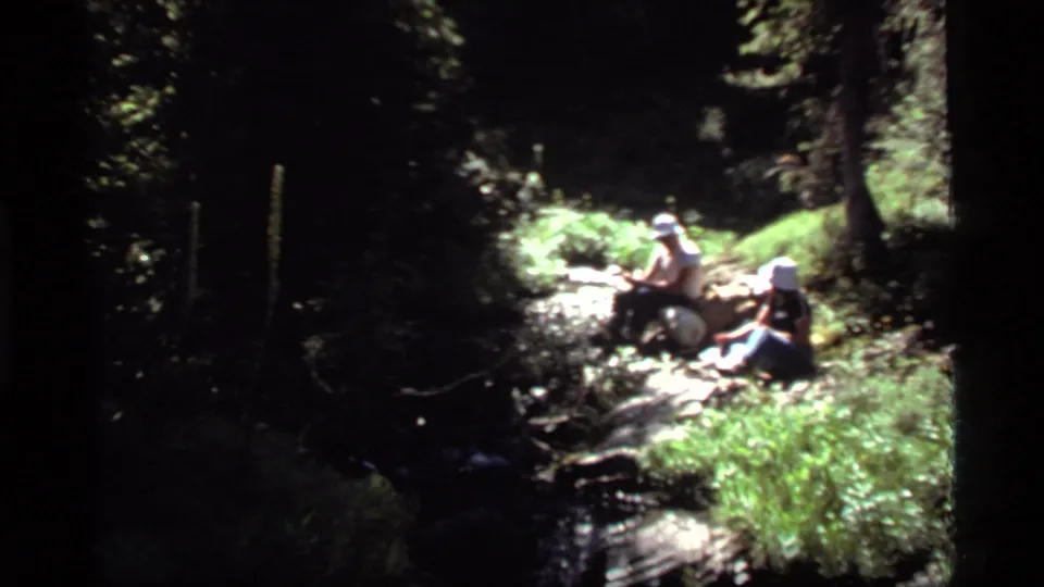 Two hikers rest beside a small stream in a wooded area near Sapphire Lake, Montana, in 1977 home-movie footage.