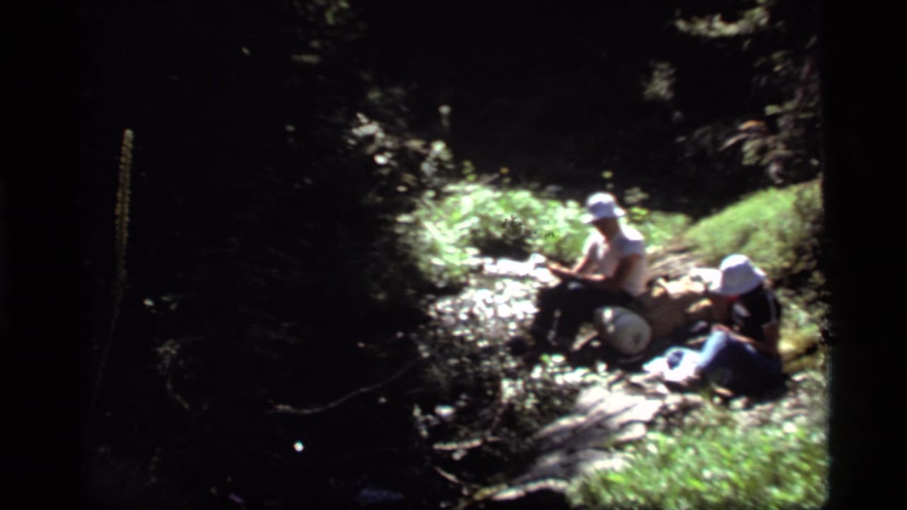 Two hikers in white hats sit beside a rocky forest stream in 1977 Sapphire Lake, Montana, home-movie footage.