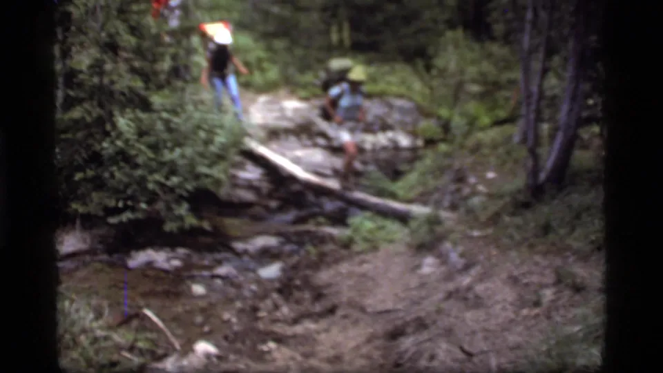 Two hikers use a fallen log to cross a small stream on a wooded trail near Sapphire Lake, Montana, in 1977 footage.