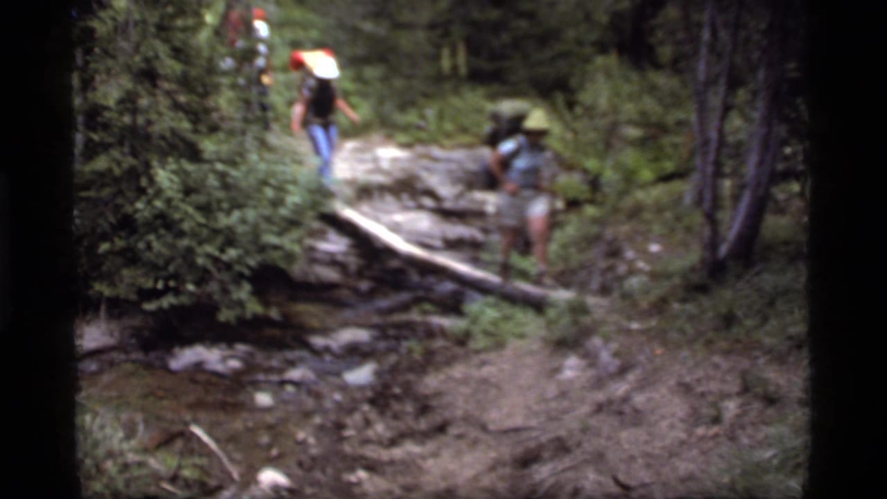 Three hikers in 1977 Sapphire Lake, Montana footage approach a fallen log across a shallow stream in dense forest.