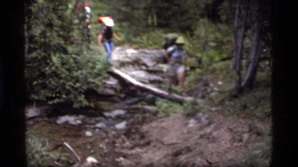 Three hikers in 1977 Sapphire Lake, Montana footage approach a fallen log across a shallow stream in dense forest.