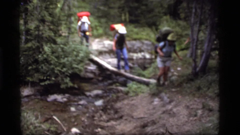 Three hikers pause and step around a log over a shallow stream in 1977 Sapphire Lake, Montana archival footage.