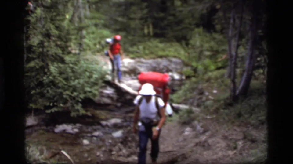 A hiker with a red backpack moves toward the camera after a log crossing in 1977 Sapphire Lake, Montana footage.