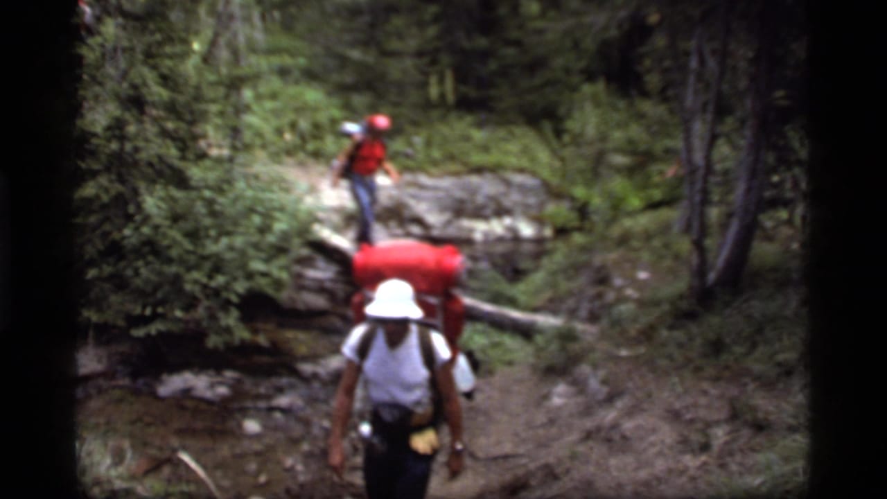 1977 8mm frame near Sapphire Lake, Montana, showing two backpackers on a wooded trail with a log over a stream.