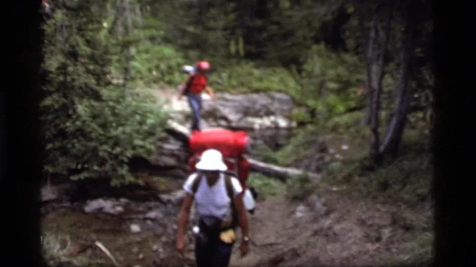 1977 8mm frame near Sapphire Lake, Montana, showing two backpackers on a wooded trail with a log over a stream.