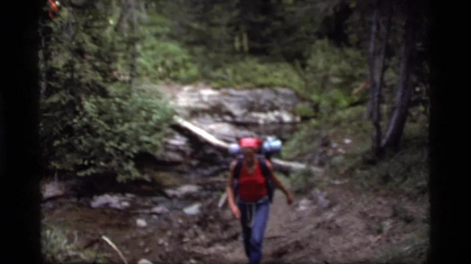 1977 home-movie frame of a backpacker in a red shirt walking on a wooded trail near Sapphire Lake, Montana.