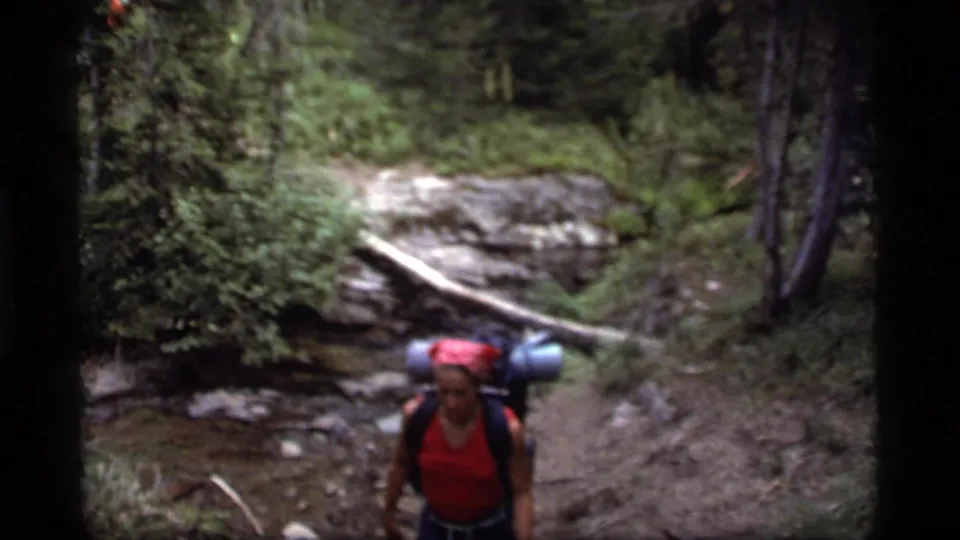 A close 8mm frame shows a backpacker with rolled gear on a wooded Sapphire Lake trail in Montana.