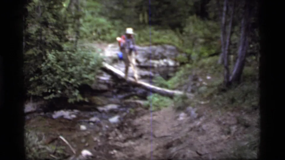 A backpacked hiker crosses a fallen log over a stream in the forest at Sapphire Lake, Montana, in 1977 home-movie footage.
