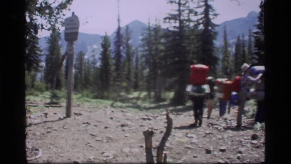 Backpackers walk away on a rocky Sapphire Lake trail, with evergreen trees, a trail sign, and distant Montana mountains visible.