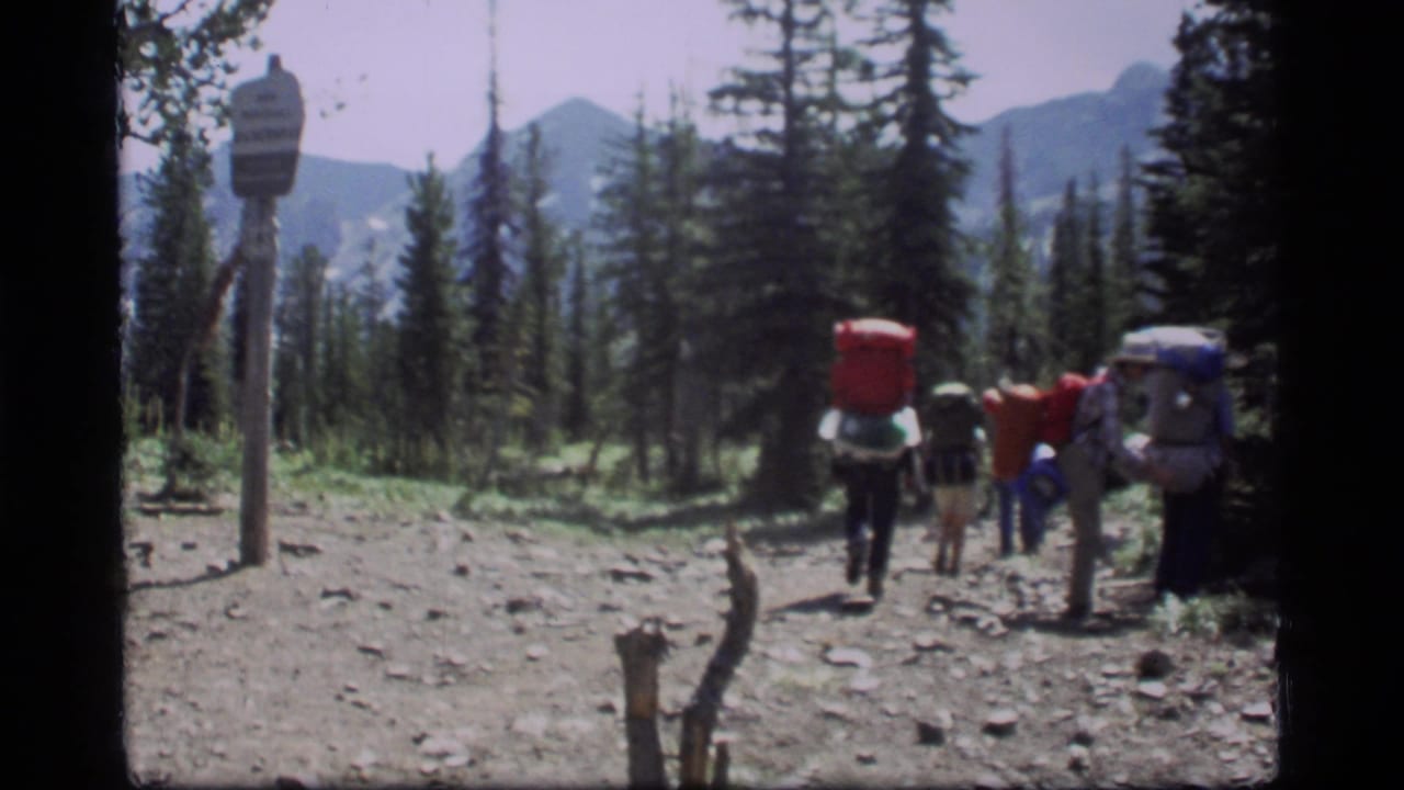 Backpackers leave a rocky forest clearing beside a wooden trail sign with mountains behind them.