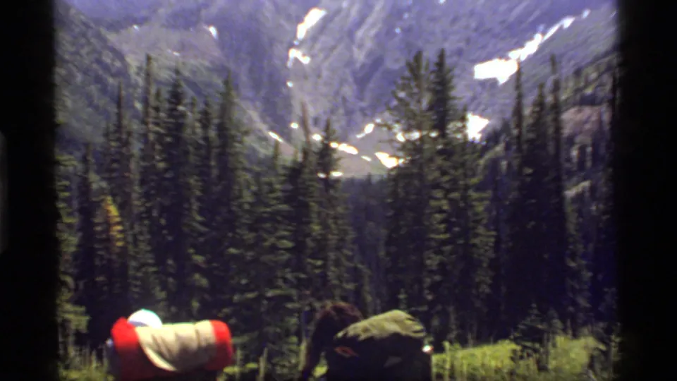 Backpackers move through forest toward a steep Montana mountain view, with snow patches on the slopes and soft 8mm exposure.