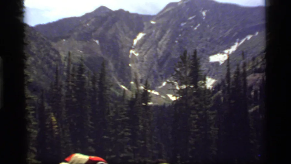 1977 Montana home-movie frame looking across evergreens toward rocky mountains with snow streaks and a cropped visitor in the foreground.