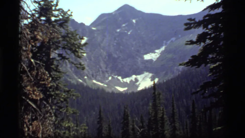 1977 Sapphire Lake, Montana frame showing a forested valley, high ridges, snow patches, and trees along the edges of the 8mm frame.