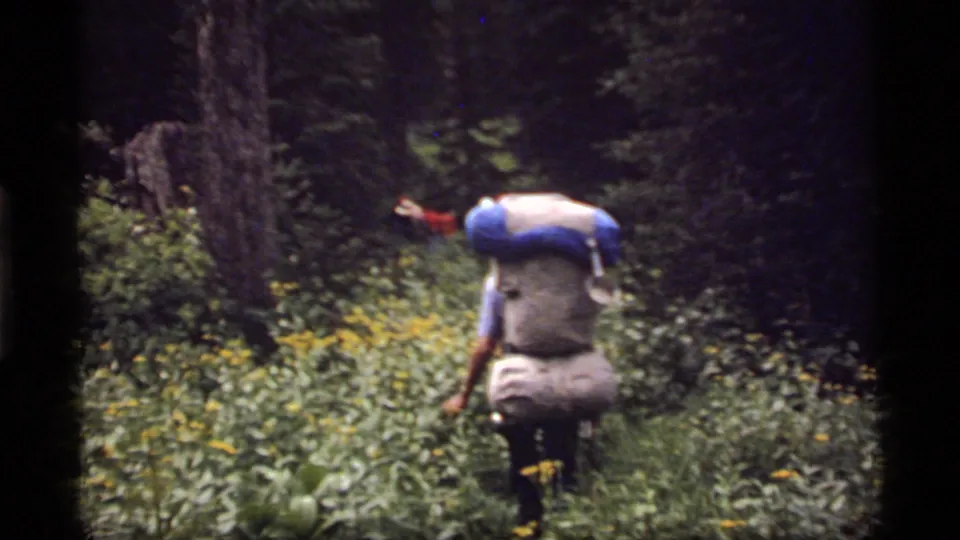 A backpacked hiker moves through a wooded Sapphire Lake, Montana trail in 1977 home-movie footage.