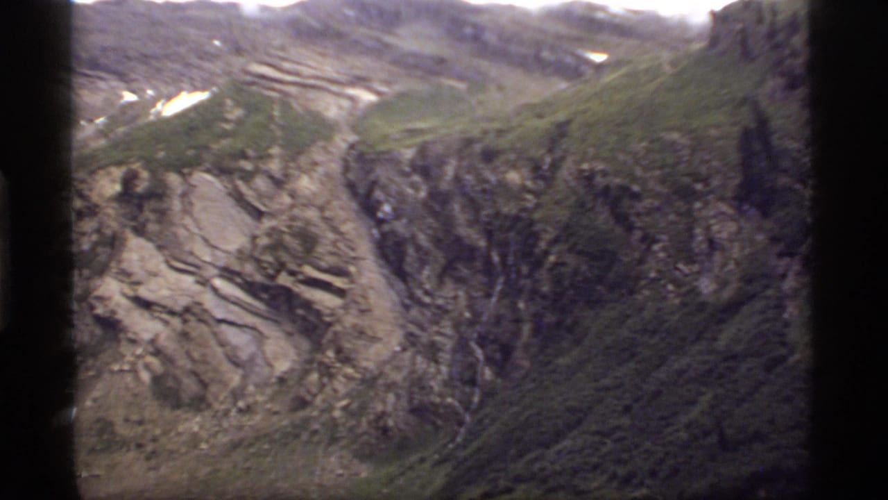 Rocky mountain slope with green vegetation, snow patches, and a narrow waterfall.