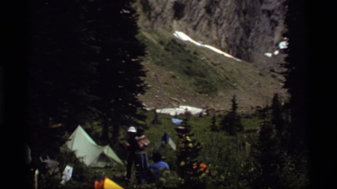 A small 1977 lakeside camp near Sapphire Lake appears among pine trees, with distant campers and tents below snow-streaked slopes.