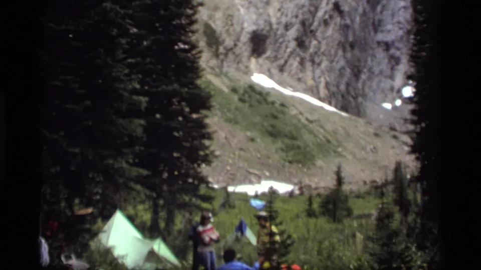 The 8mm frame shows a Montana mountain campsite with pale tents, distant people, evergreens, and lingering snow on the slope above.