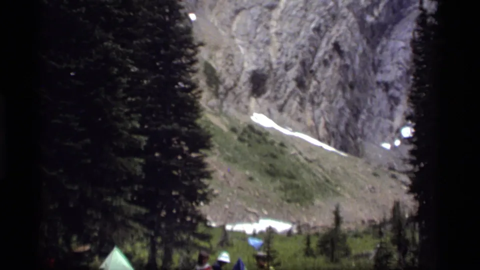 A 1977 8mm view near Sapphire Lake frames tents and campers through dark evergreens, with snow remaining on the mountain slope.