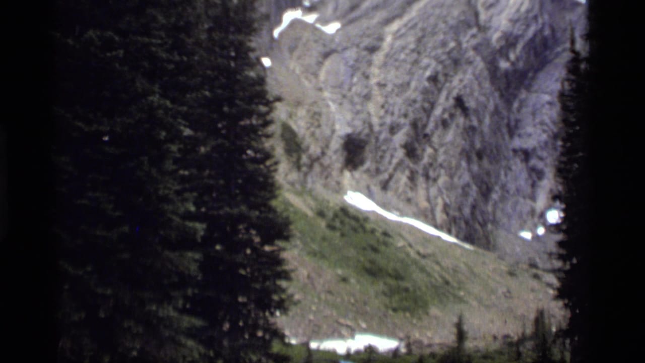 1977 8mm view near Sapphire Lake, Montana, showing dark pine trees before a rocky mountain face with lingering snow.