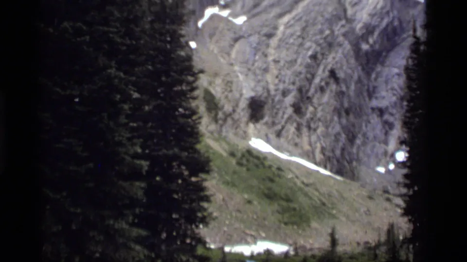 1977 8mm view near Sapphire Lake, Montana, showing dark pine trees before a rocky mountain face with lingering snow.