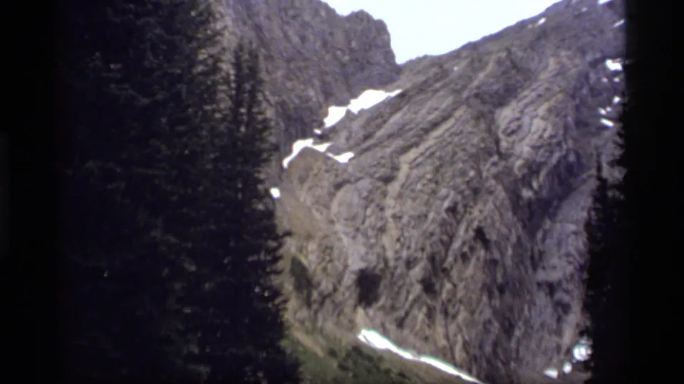 1977 Montana home-movie frame with a dark pine foreground, steep gray rock, small snowfields, and bright sky.