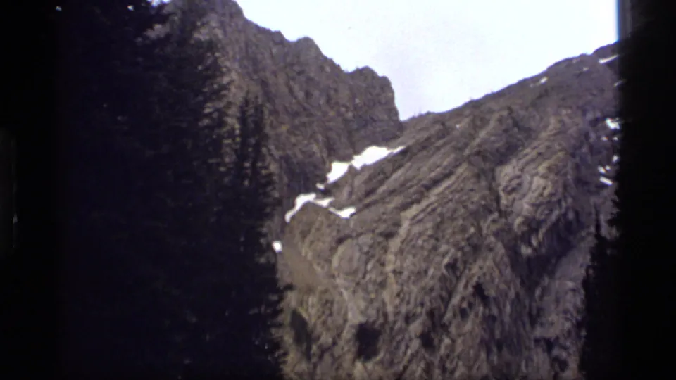 Late pan frame from 1977 Sapphire Lake footage, showing rocky ridges, scattered snow, dark pines, and pale sky.