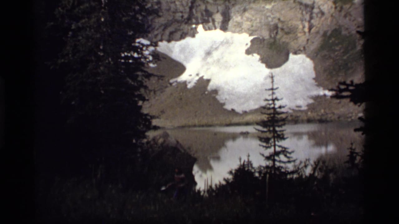 Dark evergreen foreground framing a mountain lake with snow patches reflected in still water.
