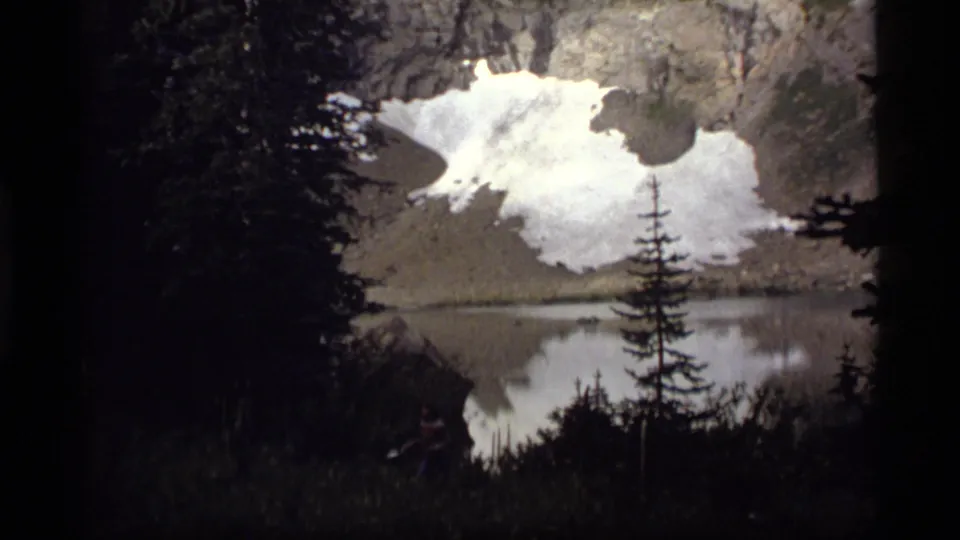 1977 Sapphire Lake, Montana footage appears to show a shadowed mountain lake with snow patches and evergreen silhouettes reflected in calm water.